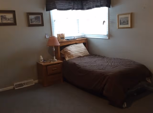 A small bedroom with a single bed covered in a brown comforter and a striped pillow. The bed is positioned under a window with blinds and a dark valance. Next to the bed is a wooden nightstand with a pink lampshade, a small white device, and some other small items. The walls are light-colored and decorated with three framed pictures.