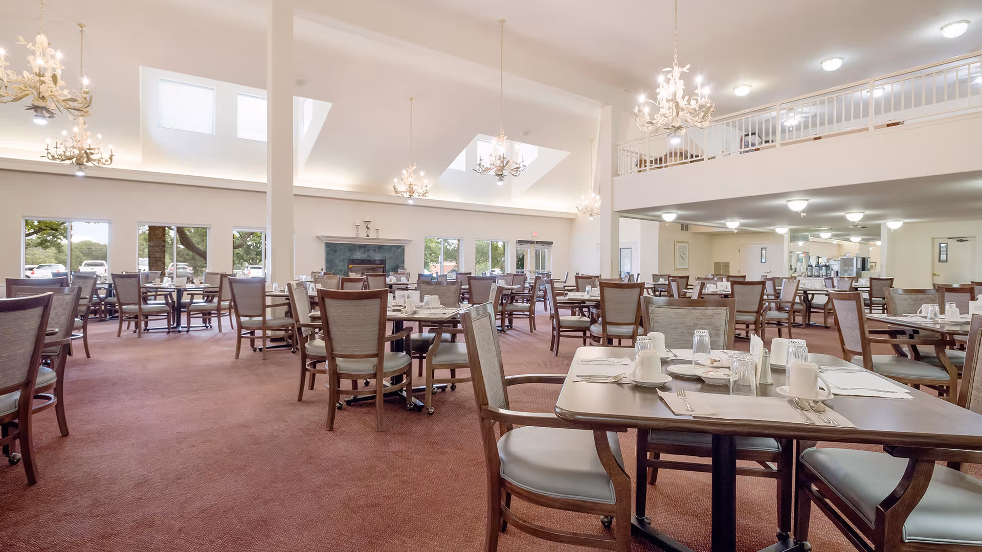 Spacious dining room with multiple wooden tables and chairs arranged neatly. Each table is set with white cups, glasses, and silverware. The room features high ceilings with chandeliers, large windows allowing natural light, and a fireplace at the far end. There is a mezzanine level with a railing overlooking the dining area.