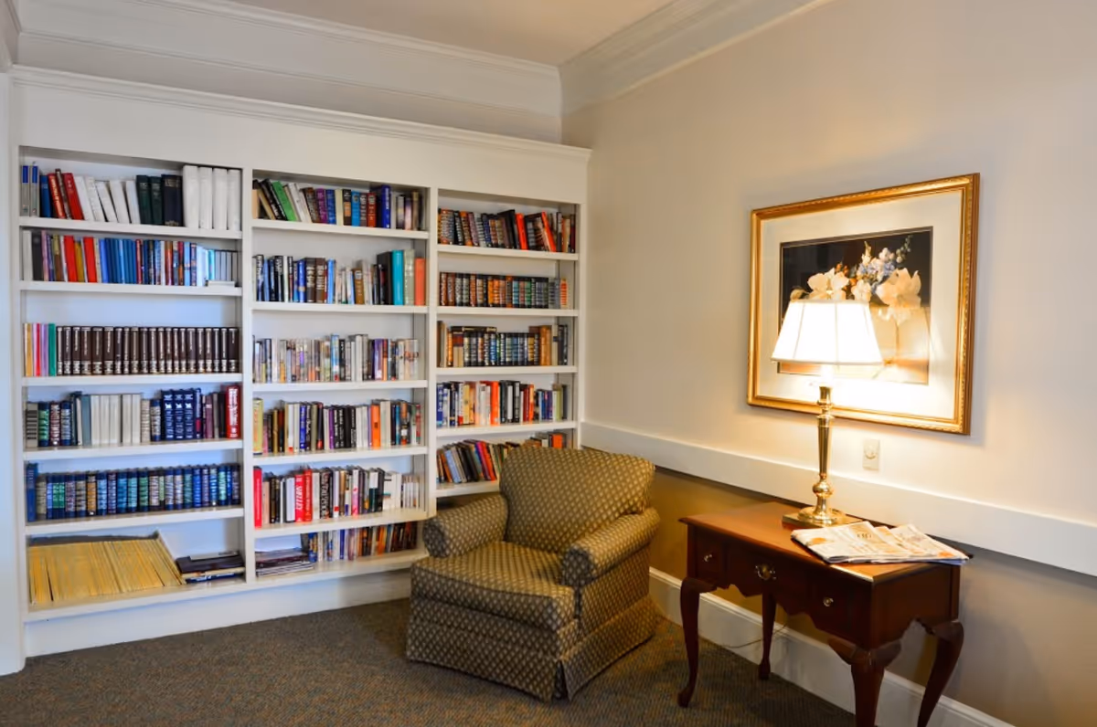 Cozy reading nook with built-in bookshelves, an upholstered armchair, a side table with a lamp and framed artwork on the wall.