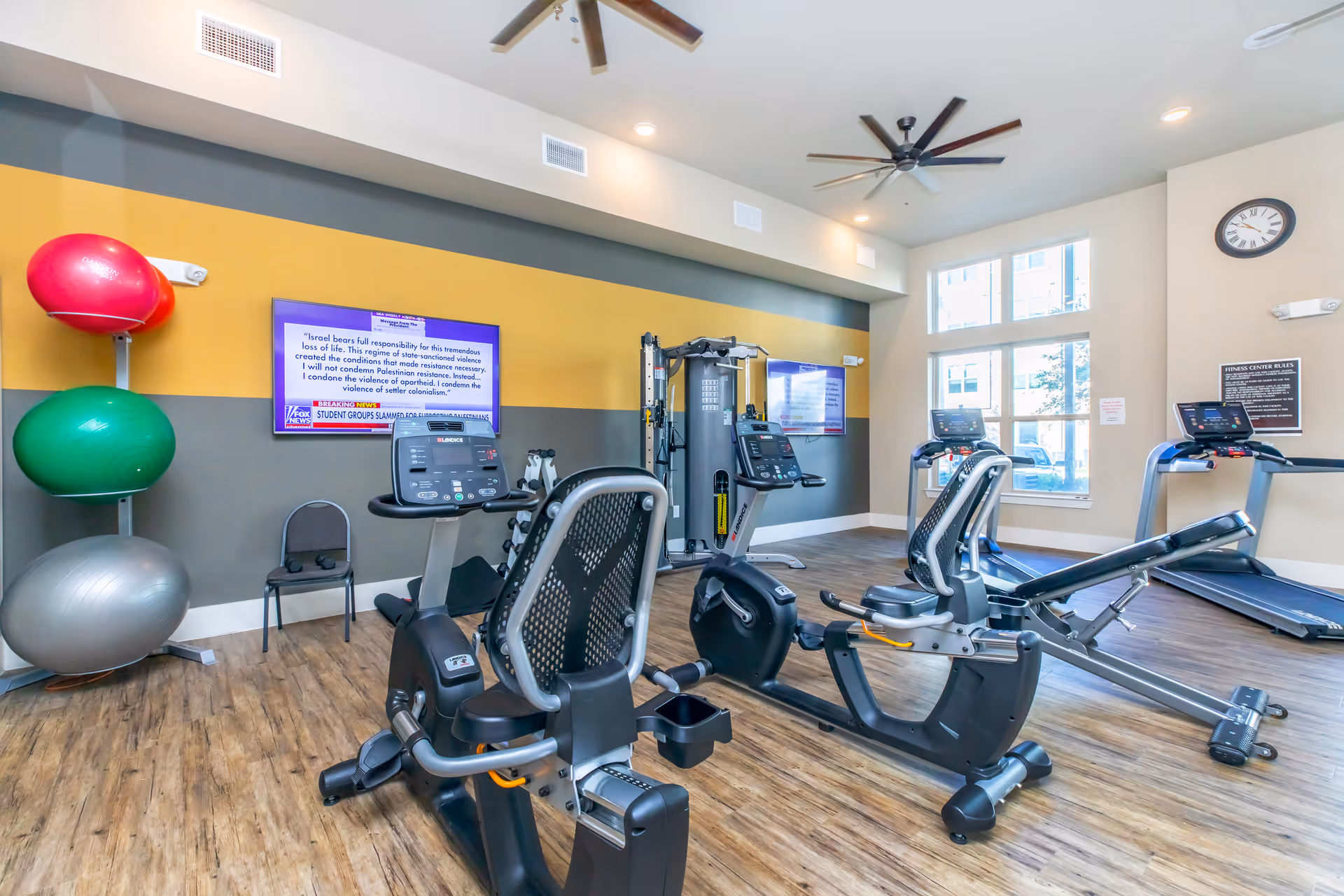 Well-lit fitness room with exercise bikes, treadmills, a multi-station weight machine, and large exercise balls against a striped wall.