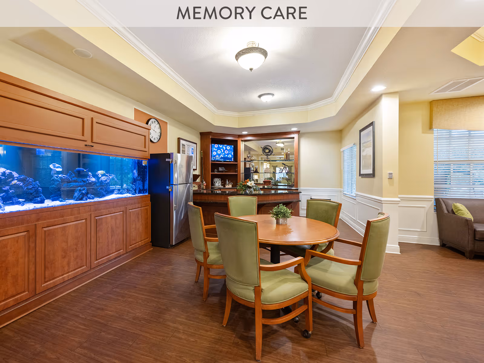 A cozy memory care room featuring a round wooden table with six green cushioned chairs on wheels. To the left, there is a large built-in aquarium with colorful fish and coral, set in wooden cabinetry. Behind the table, a small bar area with a granite countertop, a stainless steel refrigerator, and a mounted TV is visible. The room has warm yellow walls, wood flooring, and a comfortable seating area with a brown sofa near windows with blinds.