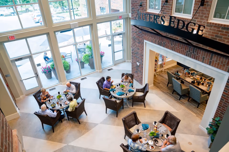 Overhead view of seniors eating at round tables in a bright communal dining area with large windows and a 'RIVER'S EDGE' sign on the wall.