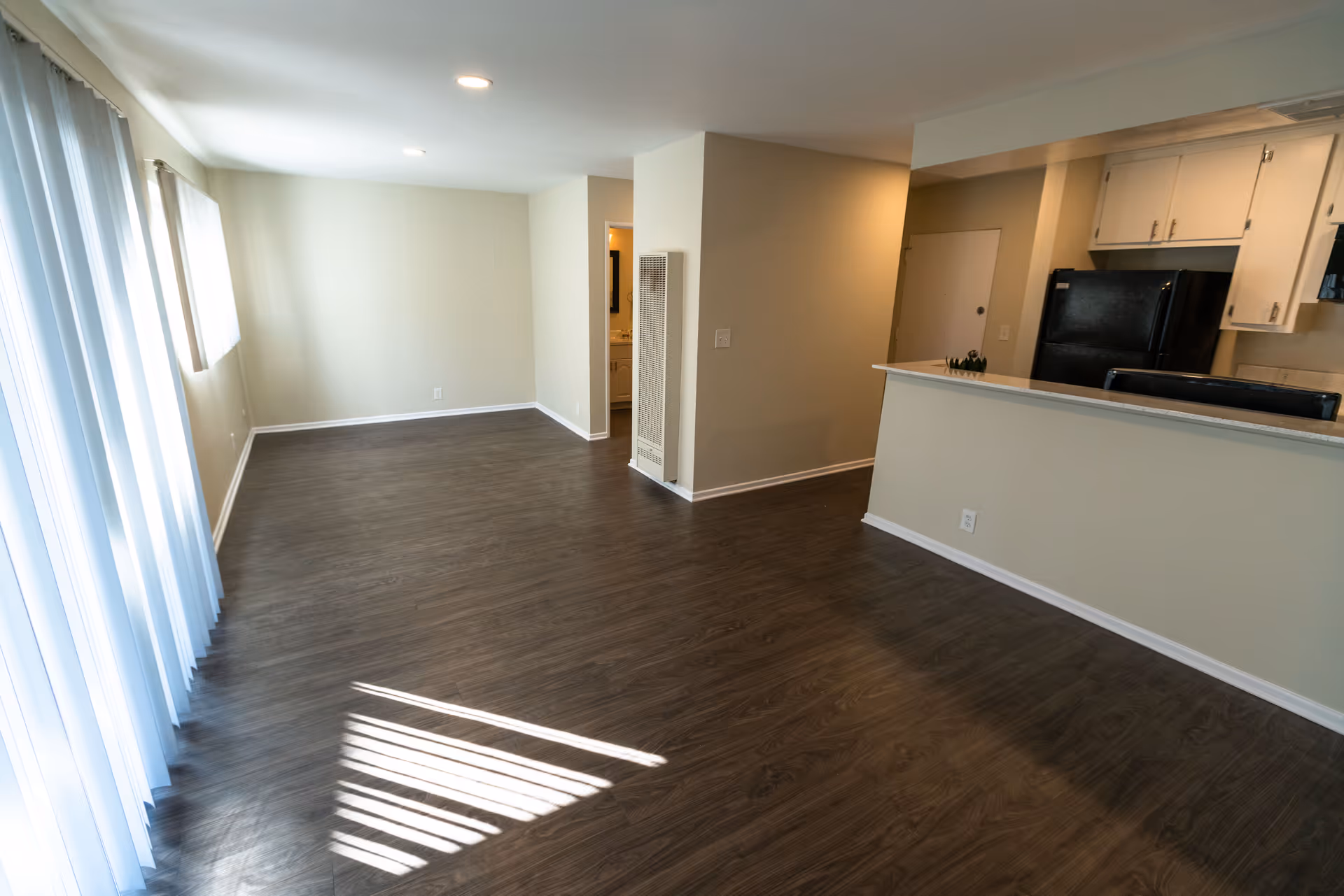 Empty interior space with wood flooring, beige walls, recessed ceiling lights, and vertical blinds covering a large window. The space includes a kitchen area with white cabinets and a black refrigerator, and an open doorway leading to another room.