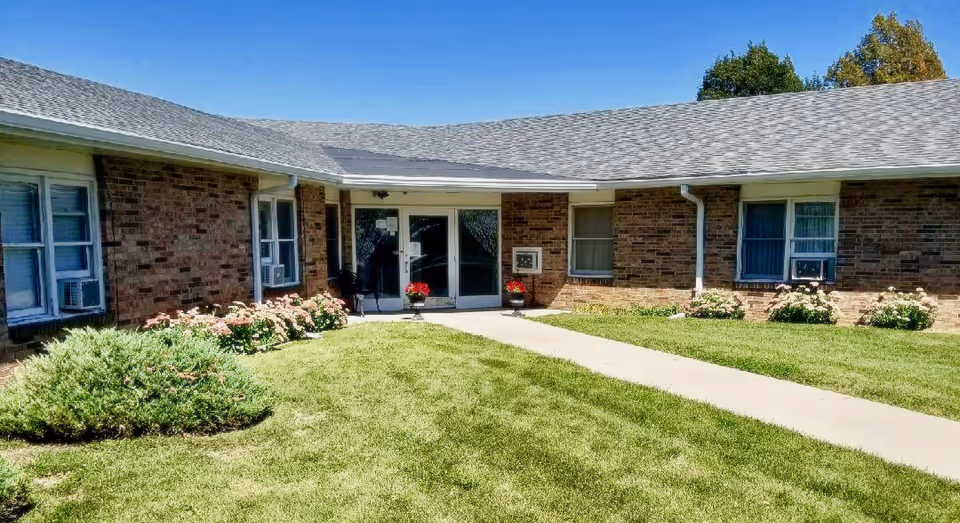 Single-story brick senior living facility entrance with a walkway, lawn, flower beds, and glass double doors.