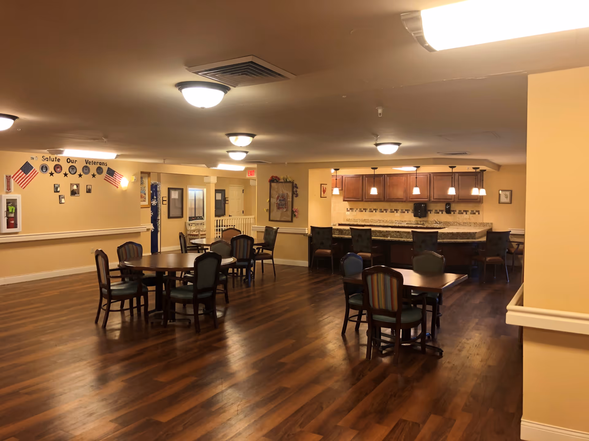 Interior view of a senior living facility dining area with several wooden tables and chairs arranged on a polished wooden floor. In the background, there is a counter with bar stools and hanging pendant lights. The walls are painted a warm beige color, and there is a display on the left wall with American flags and a 'Salute Our Veterans' sign.