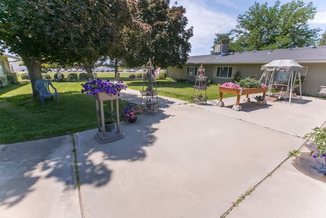 Outdoor patio area at River's Edge Rehabilitation & Living Center featuring a concrete walkway, green lawn with trees, several flower pots with purple flowers, garden trellises, a wooden planter box with pink flowers, a swing bench with a canopy, and a single plastic chair under a tree.