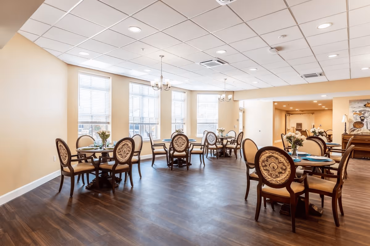 A bright dining room with multiple round wooden tables, each surrounded by four upholstered chairs with ornate backs. The room has large windows with white blinds, beige walls, dark wood flooring, and ceiling lights including chandeliers. There are floral centerpieces on each table and a sideboard with a painting and lamp in the background.