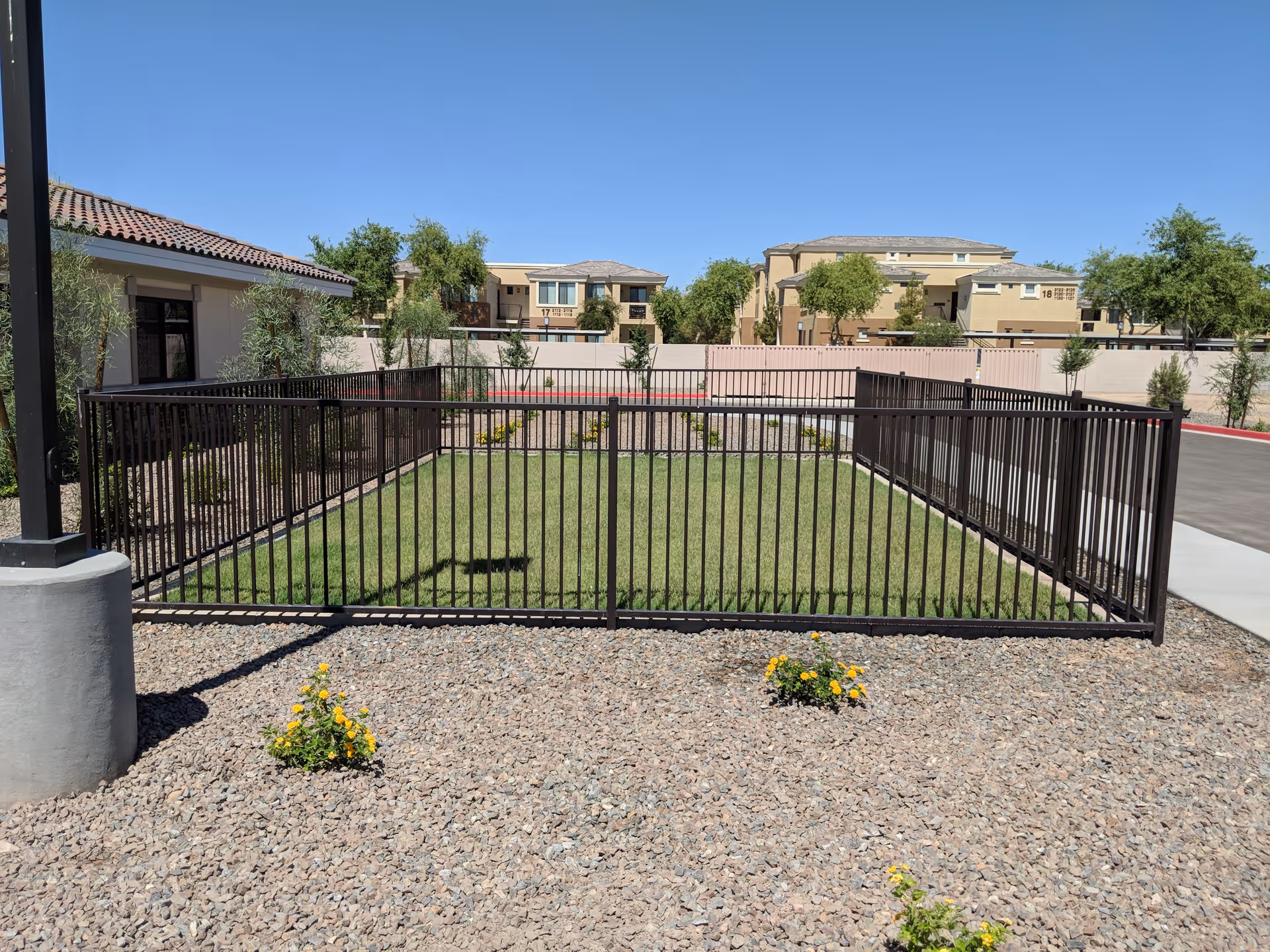 A fenced grassy area with a black metal fence surrounding it, located outdoors near a building with a tiled roof. There are small yellow flowers planted in the gravel in front of the fence, and residential buildings and trees are visible in the background under a clear blue sky.