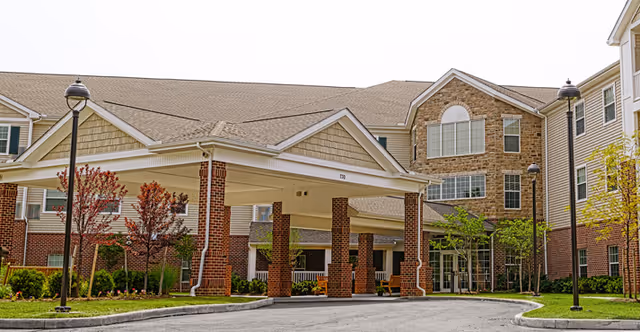 Front exterior view of Danbury Westerville senior living facility showing a covered entrance with brick pillars, surrounding landscaping with small trees and bushes, and a multi-story building with beige siding and stone accents.