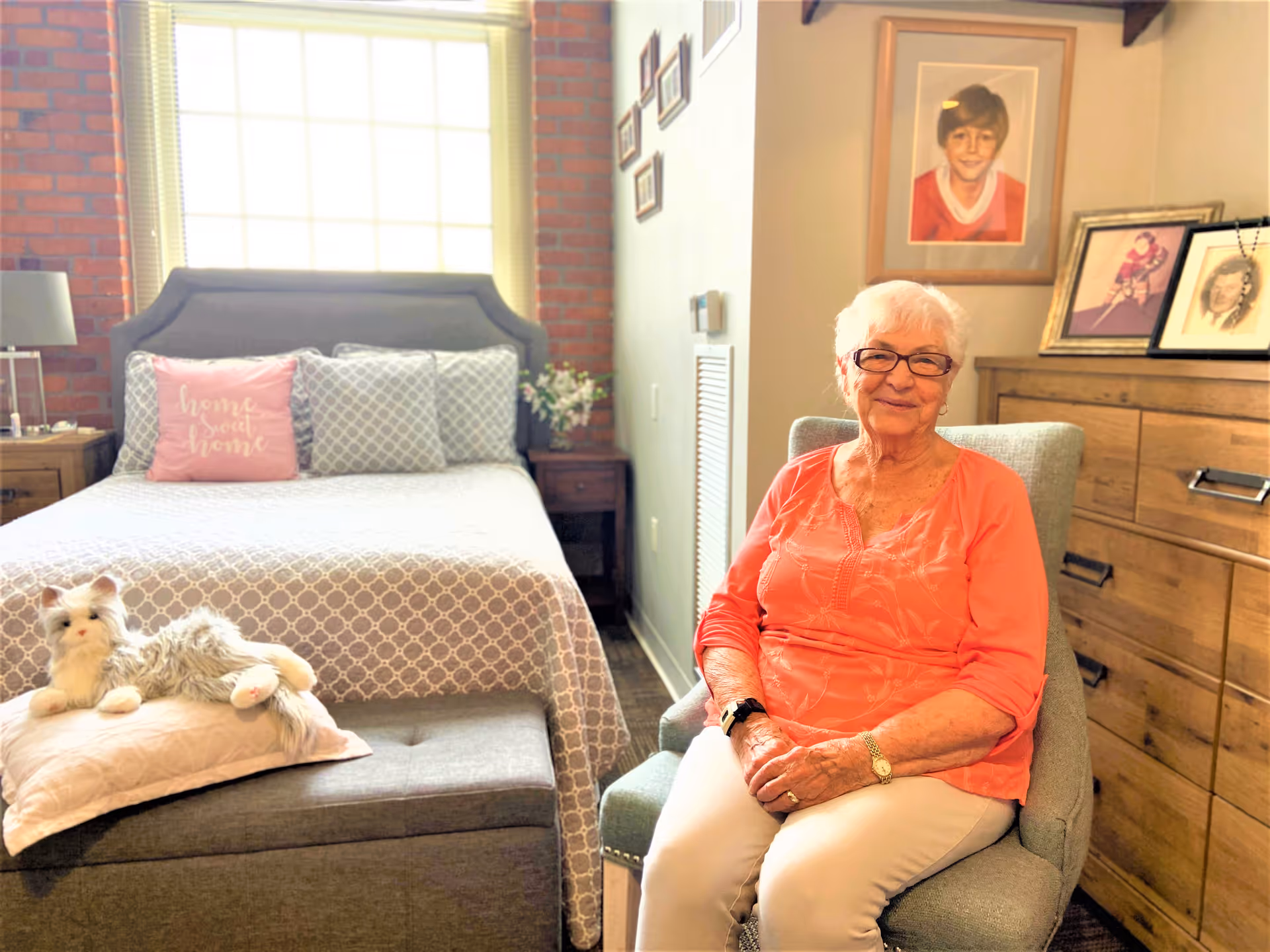 An elderly woman with white hair and glasses sits smiling in a light green armchair in a cozy bedroom. The room features a bed with a gray patterned bedspread and pillows, including a pink pillow with the words 'home sweet home'. A stuffed cat toy lies on a pillow at the foot of the bed. There is a wooden dresser with framed photos and portraits on top, and a window with blinds letting in natural light behind the bed.