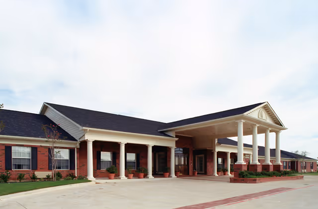 Exterior view of a single-story brick building with white columns and a covered entrance, surrounded by a paved driveway and some landscaping with small plants and shrubs.