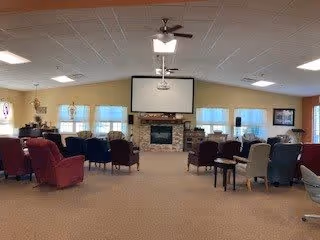 A spacious living room area with multiple chairs arranged in rows facing a stone fireplace and a large screen mounted above it. The room has beige walls, several windows with blinds, ceiling lights, and a ceiling fan. There are small tables between some chairs and framed pictures on the walls.