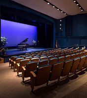 Empty auditorium with rows of seats facing a blue-lit stage featuring a grand piano.
