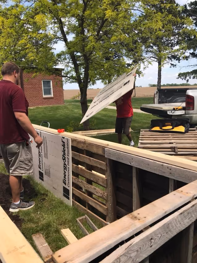 Two men working outdoors on a wooden structure, one holding a large panel labeled 'EnergyShield Continuous Wall Insulation'. They are surrounded by green grass, trees, and a brick building in the background. A pickup truck with tools is parked nearby.