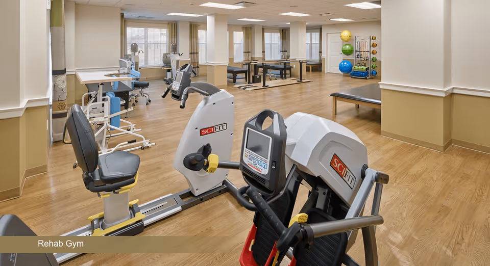 A spacious rehab gym with exercise equipment including SciFit machines, therapy tables, and various exercise balls and equipment organized along the walls. The room has wooden flooring, beige walls, and large windows allowing natural light to enter.