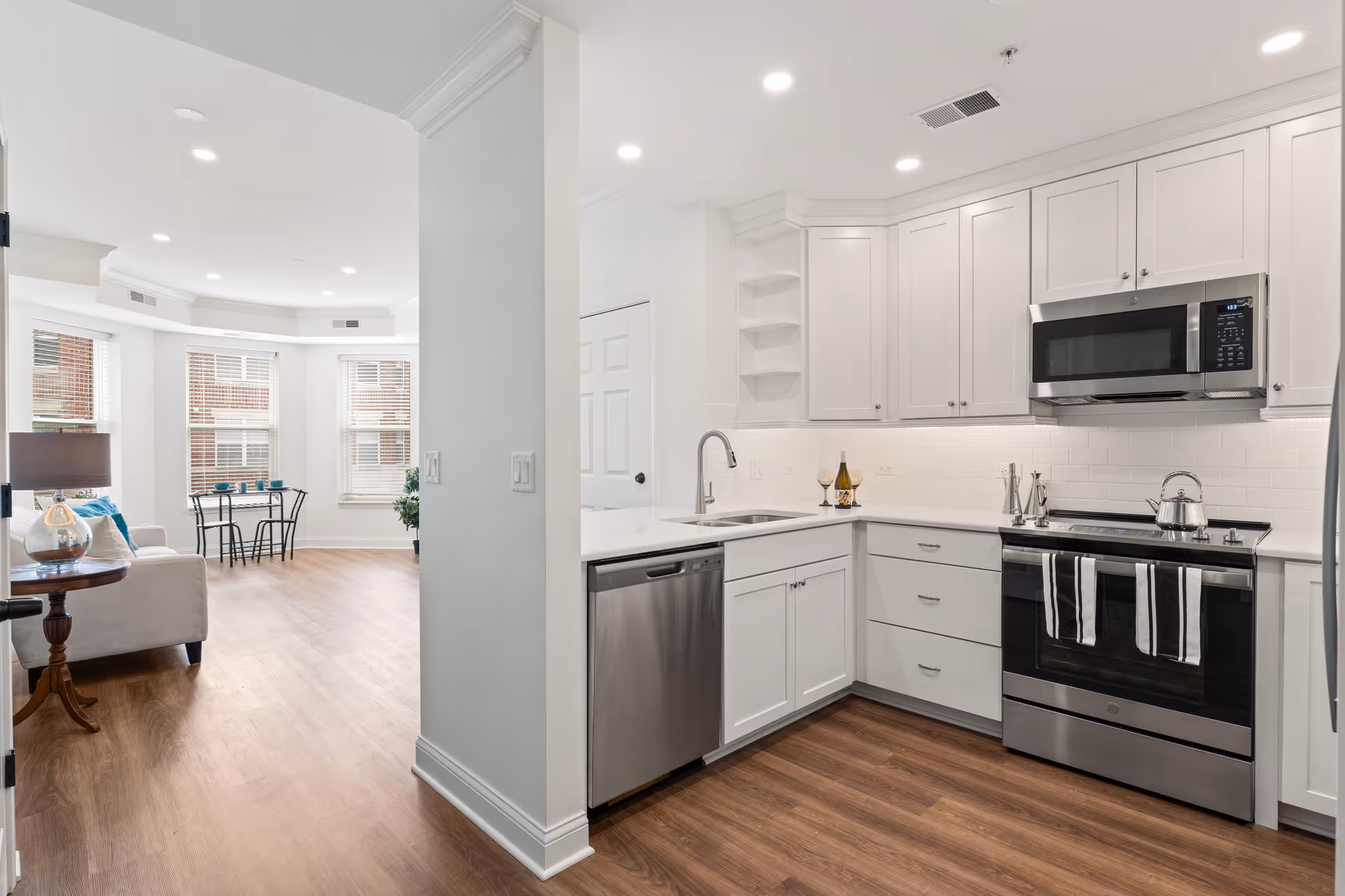 Bright and modern kitchen with white cabinets, stainless steel appliances including a dishwasher, oven, and microwave, and a white countertop. Adjacent to the kitchen is a living area with a small round table and two chairs near three large windows with blinds, and a beige sofa with a side table and lamp.