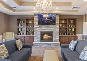 A cozy living room featuring two dark gray sofas with patterned pillows facing each other across a beige ottoman. In the center is a stone fireplace with a lit fire and a flat-screen TV mounted above it. Built-in wooden shelves flank the fireplace, decorated with various vases and ornaments. A chandelier hangs from the ceiling, illuminating the room warmly.