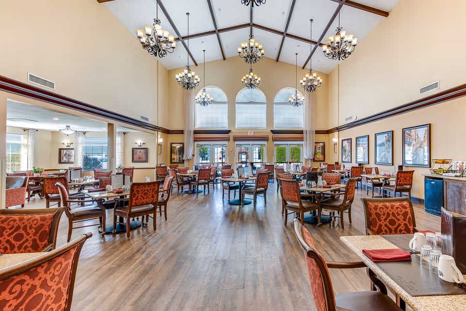 A spacious and well-lit dining room with high vaulted ceilings and multiple chandeliers. The room features several tables with red patterned chairs arranged neatly on a wooden floor. Large windows and glass doors at the far end allow natural light to fill the space. The walls are decorated with framed artwork and there is a sideboard with cups and other dining essentials.