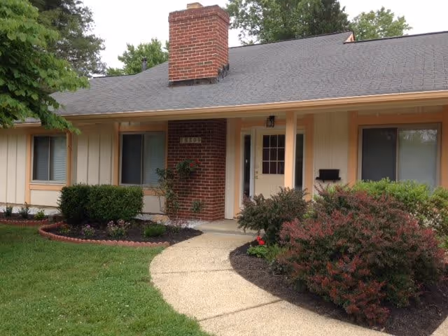 Front exterior view of a single-story residential building with a brick chimney, beige siding, two windows, a white front door, and a curved concrete walkway leading to the entrance. The yard has green grass, bushes, and small flower beds.