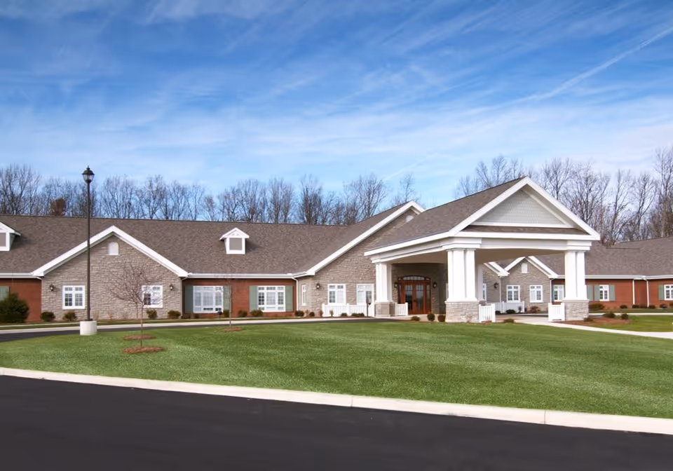 Front exterior of a single-story brick and stone senior living building with a large covered entrance, manicured lawn, and clear blue sky.