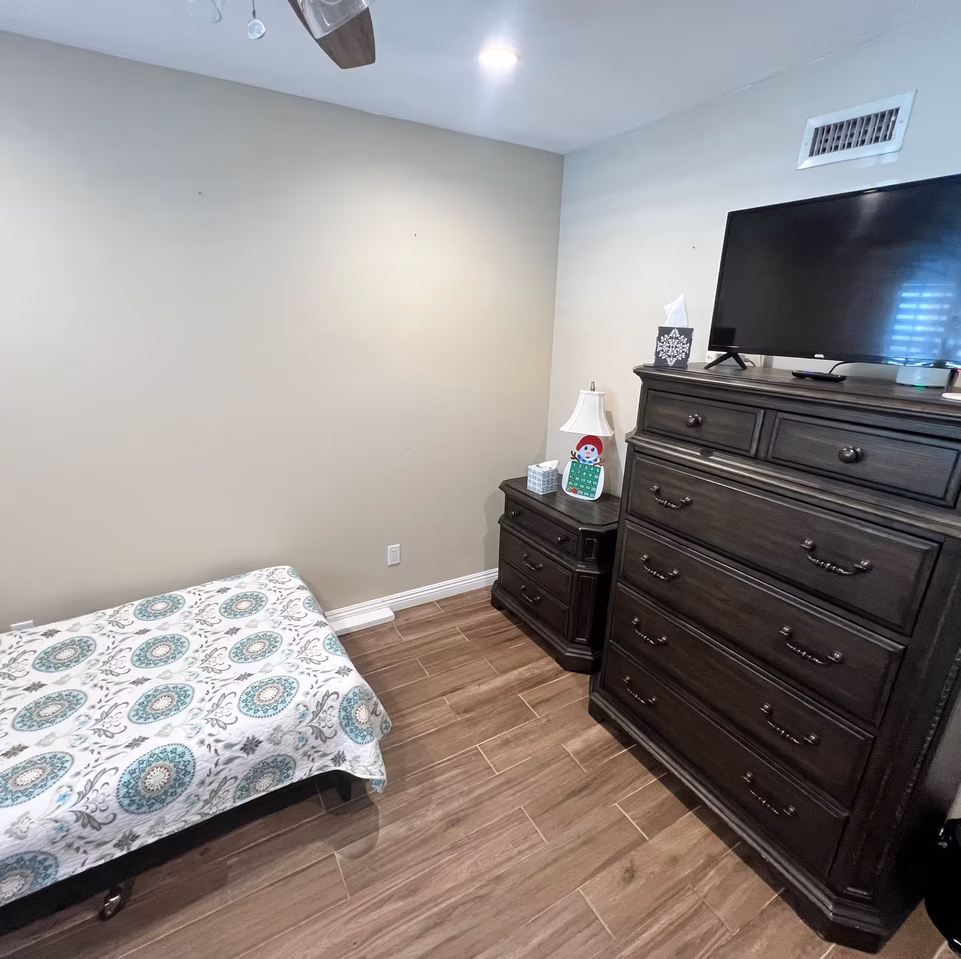 Small bedroom with a single bed covered in patterned bedding, a dark wooden dresser and nightstand with a lamp and TV on top.