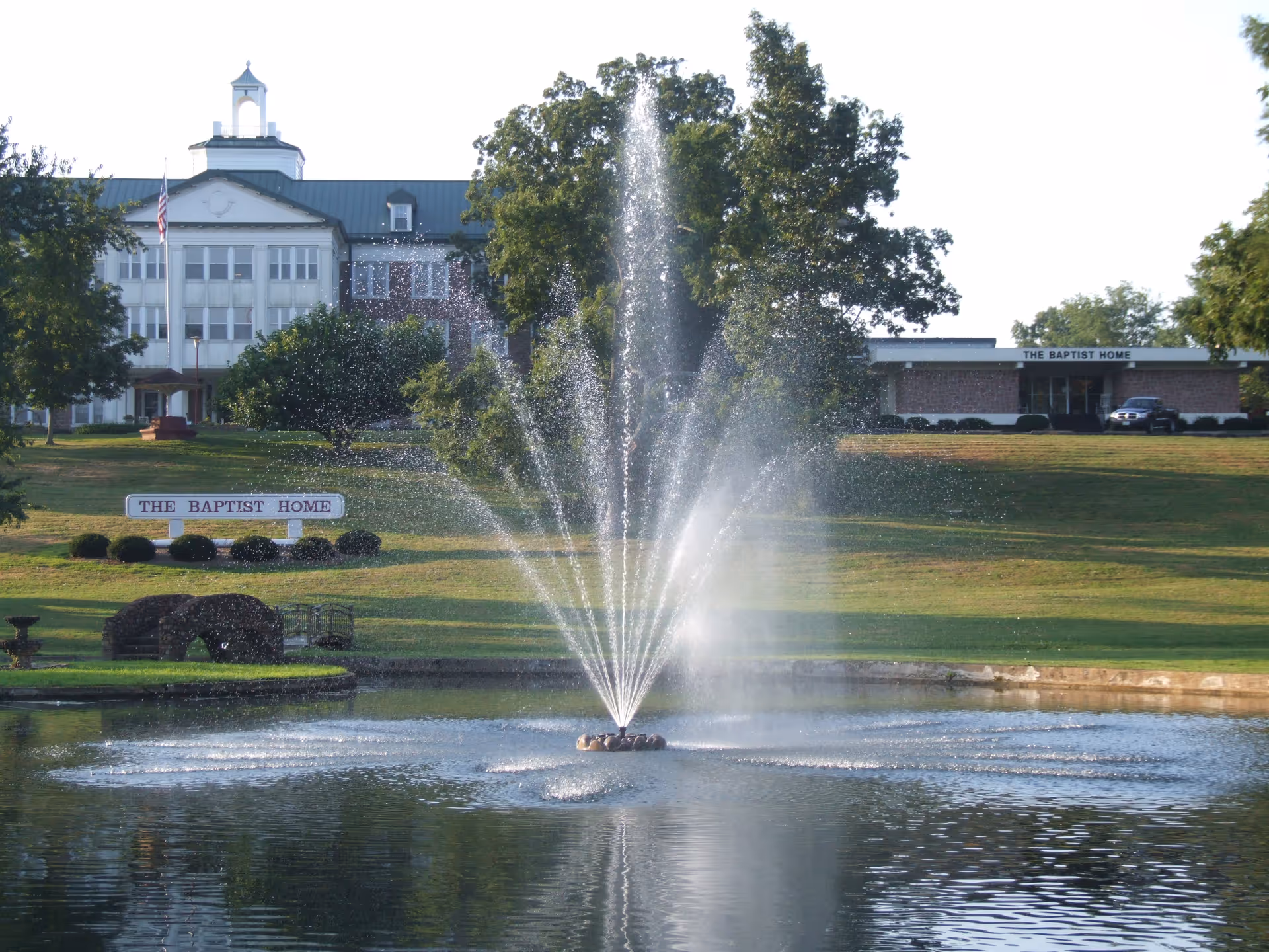 A large water fountain sprays water in the middle of a pond with a grassy area and trees in the background. Behind the pond, there are two buildings with signs reading 'THE BAPTIST HOME'. The sky is clear and the scene is well-lit by daylight.