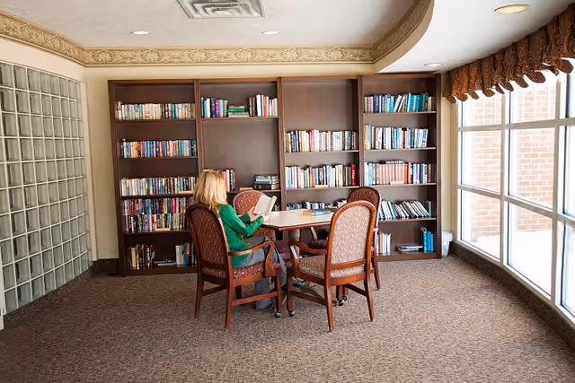 A woman with blonde hair wearing a green sweater is sitting at a round table reading a book in a quiet room with bookshelves filled with books. The room has carpeted floors, large windows with brown curtains, and a wall made of glass blocks.