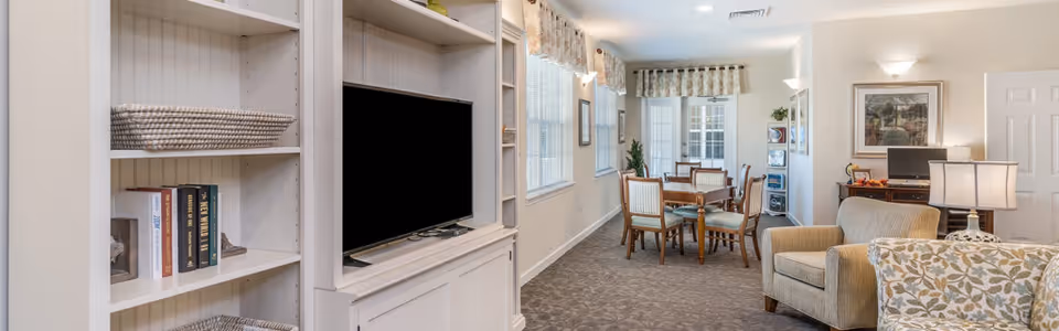 Bright communal living area with built-in white shelving and a TV, upholstered armchairs, and a dining table near windows.