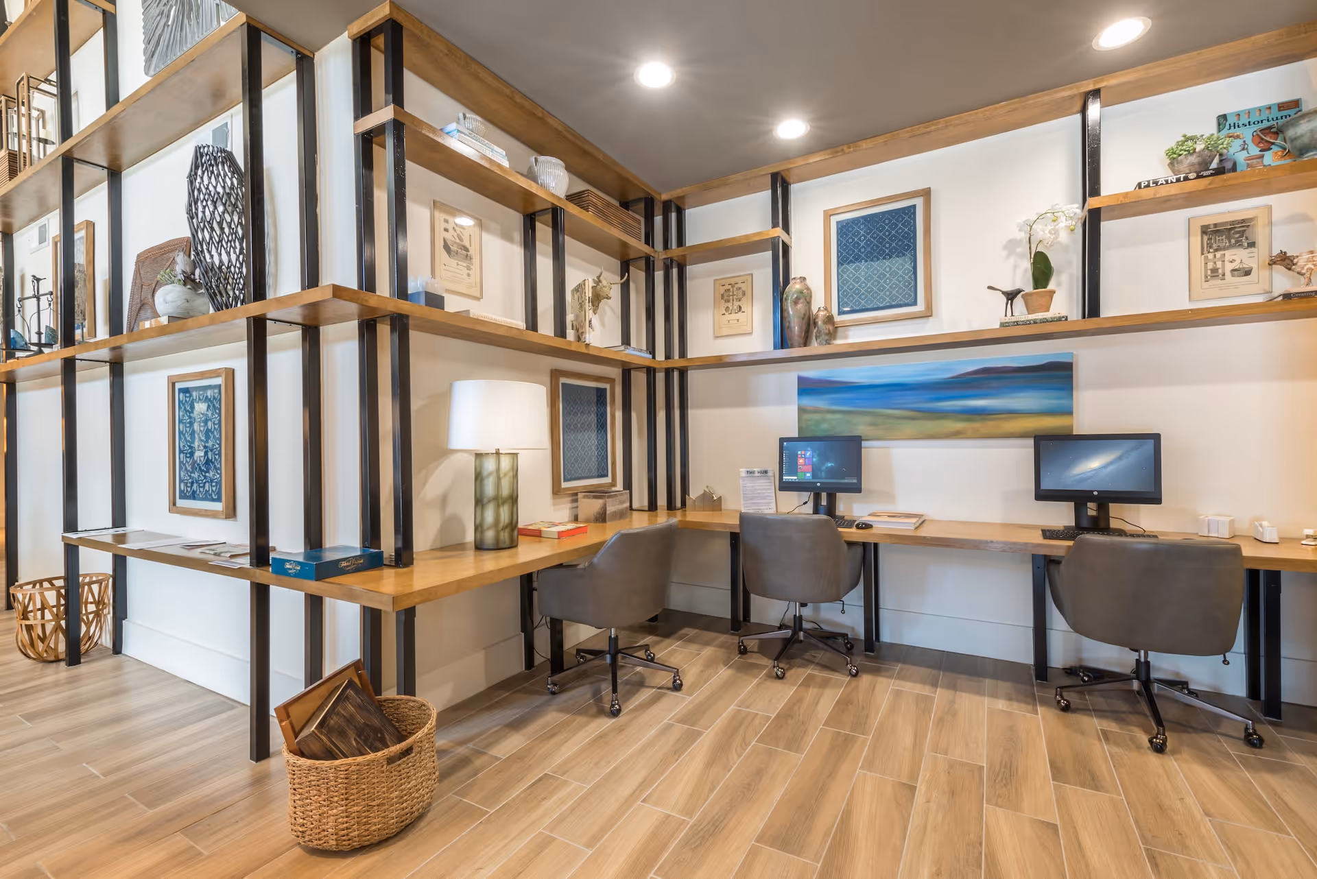 A modern workspace area with two computer desks and chairs, surrounded by wooden shelves holding decorative items, books, and framed artwork. The room has wood-look tile flooring and recessed ceiling lights.