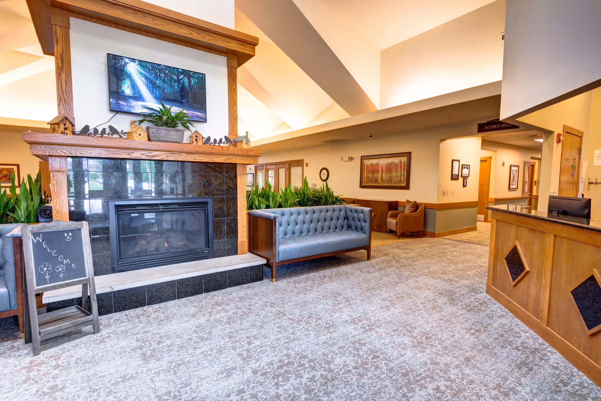 Spacious senior living facility lobby with a fireplace, tufted sofas, a reception desk, and decorative plants.