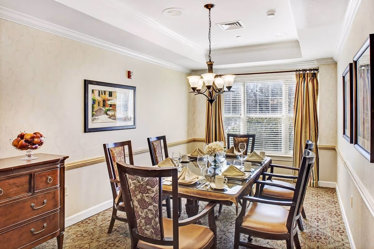 A well-lit dining room with a rectangular wooden table set for six people. The table is arranged with folded napkins, glasses, cups, and a floral centerpiece. The room features patterned carpet, beige walls with a chair rail, a wooden sideboard with a bowl of fruit, framed artwork on the walls, and a large window with striped curtains allowing natural light to enter. A chandelier hangs above the table.