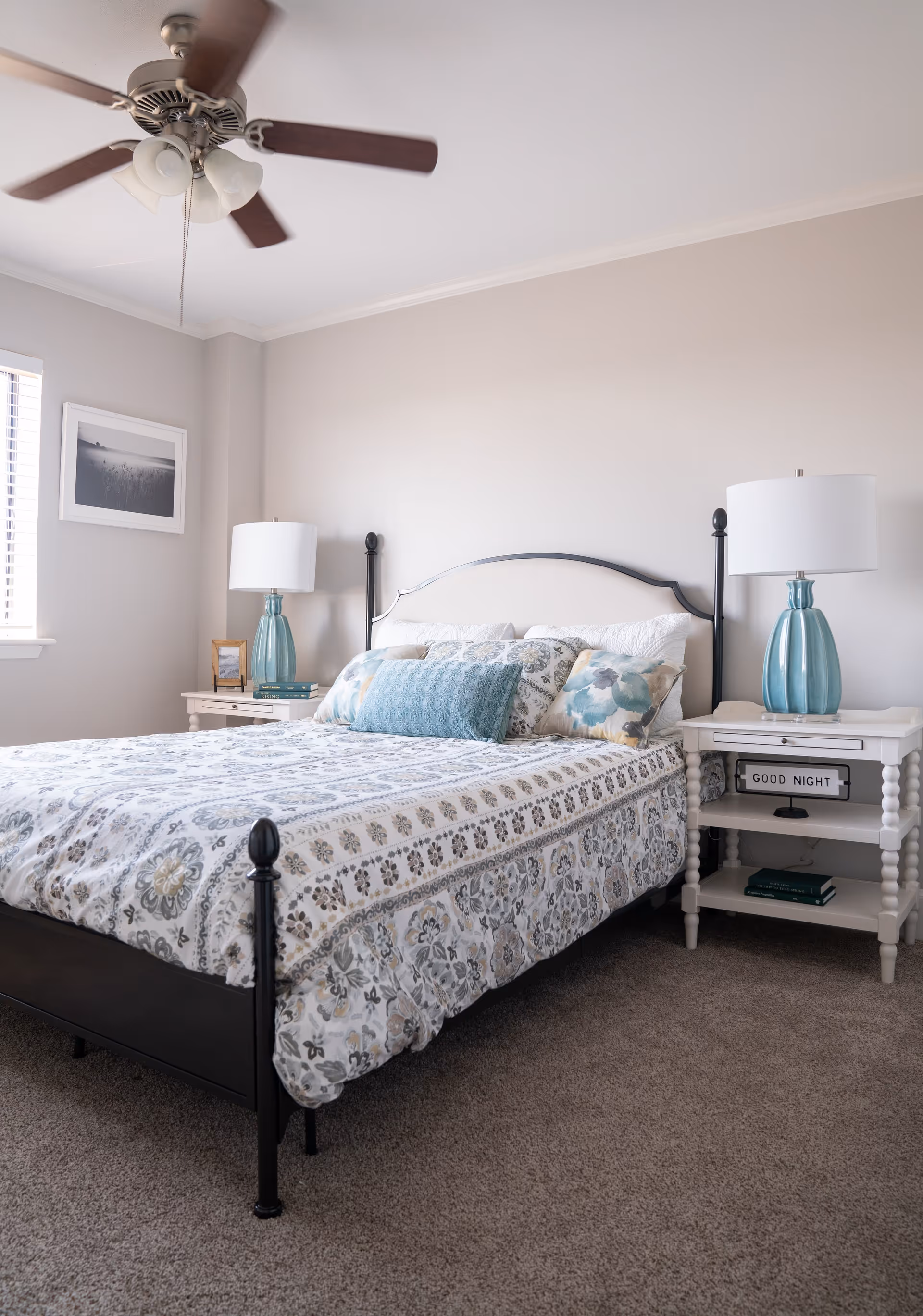 A neatly made bed with a black metal frame and patterned bedding in a bright bedroom. There are two white nightstands on either side of the bed, each with a blue glass lamp. One nightstand has a small sign that reads 'GOOD NIGHT'. A ceiling fan with lights is visible above the bed, and a window with blinds and a framed picture are on the left wall.