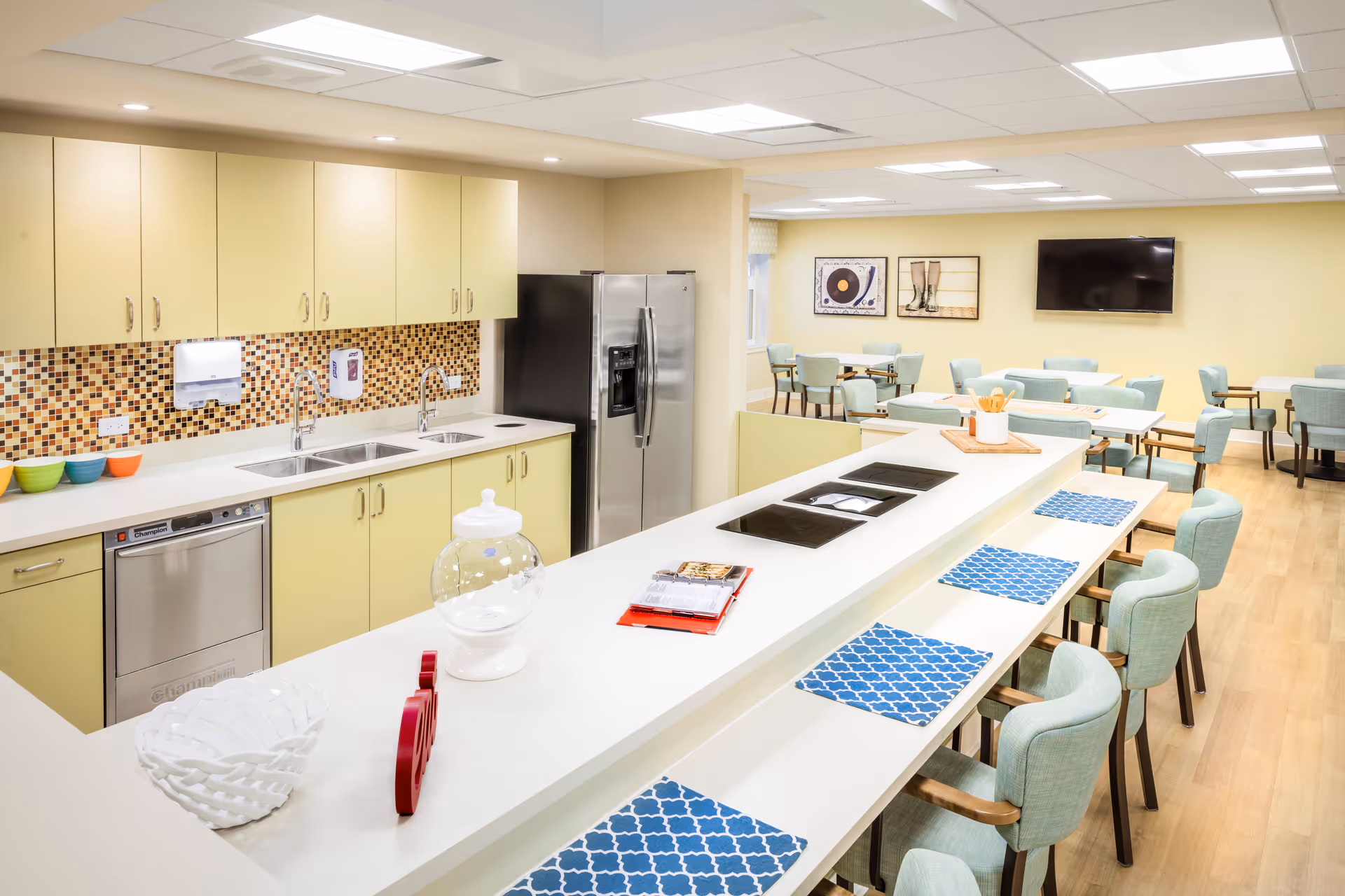 A bright and modern kitchen and dining area in a senior living facility. The kitchen features light yellow cabinets, a mosaic tile backsplash, double sinks, a dishwasher, and a stainless steel refrigerator. In the foreground, there is a long white counter with blue patterned placemats and light green cushioned chairs. In the background, there are several tables and chairs arranged for dining, a wall-mounted TV, and framed artwork on the walls.