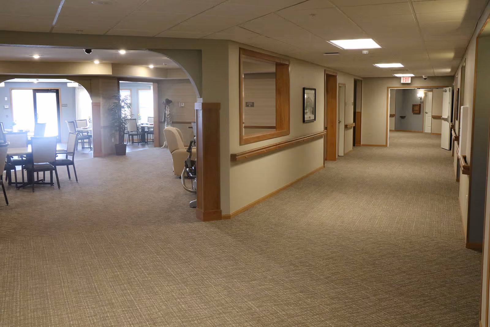 Carpeted hallway in a senior living facility with handrails and doors on the right and a dining/lounge area visible to the left.