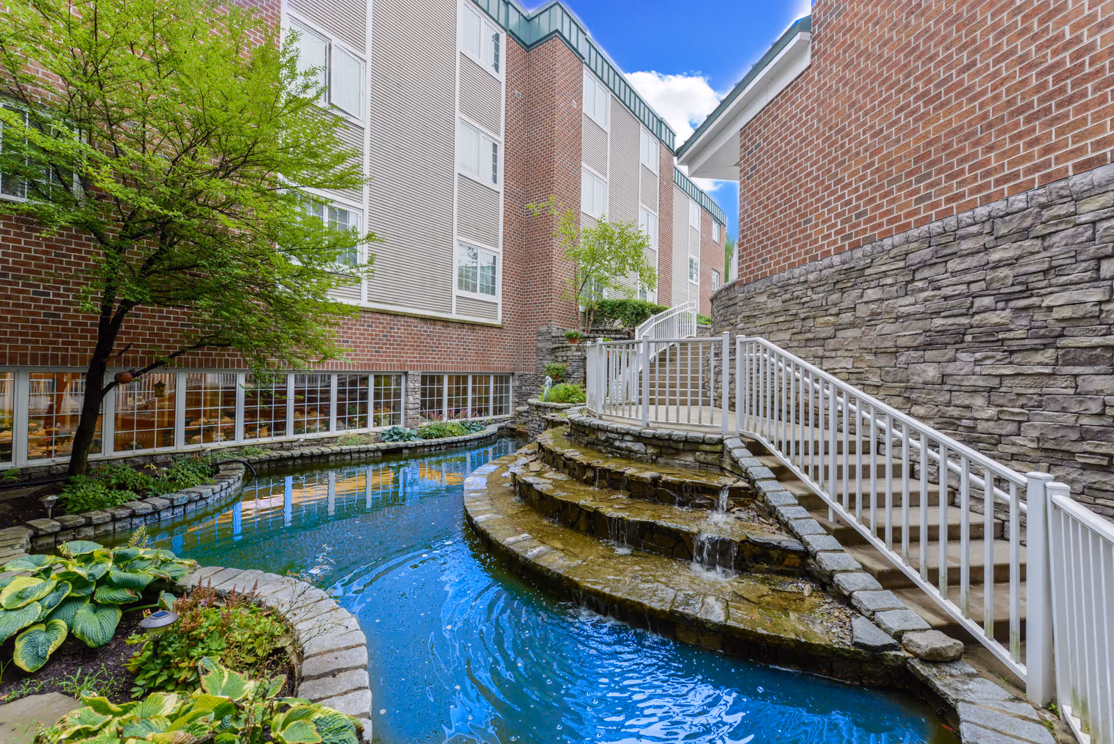 Outdoor courtyard area at Bethel Health Care Center featuring a small cascading waterfall flowing into a narrow pond surrounded by stone edging and greenery. There are brick and stone building walls with multiple windows and a white railing along a staircase leading up beside the waterfall. A tree with green leaves is visible on the left side under a partly cloudy blue sky.
