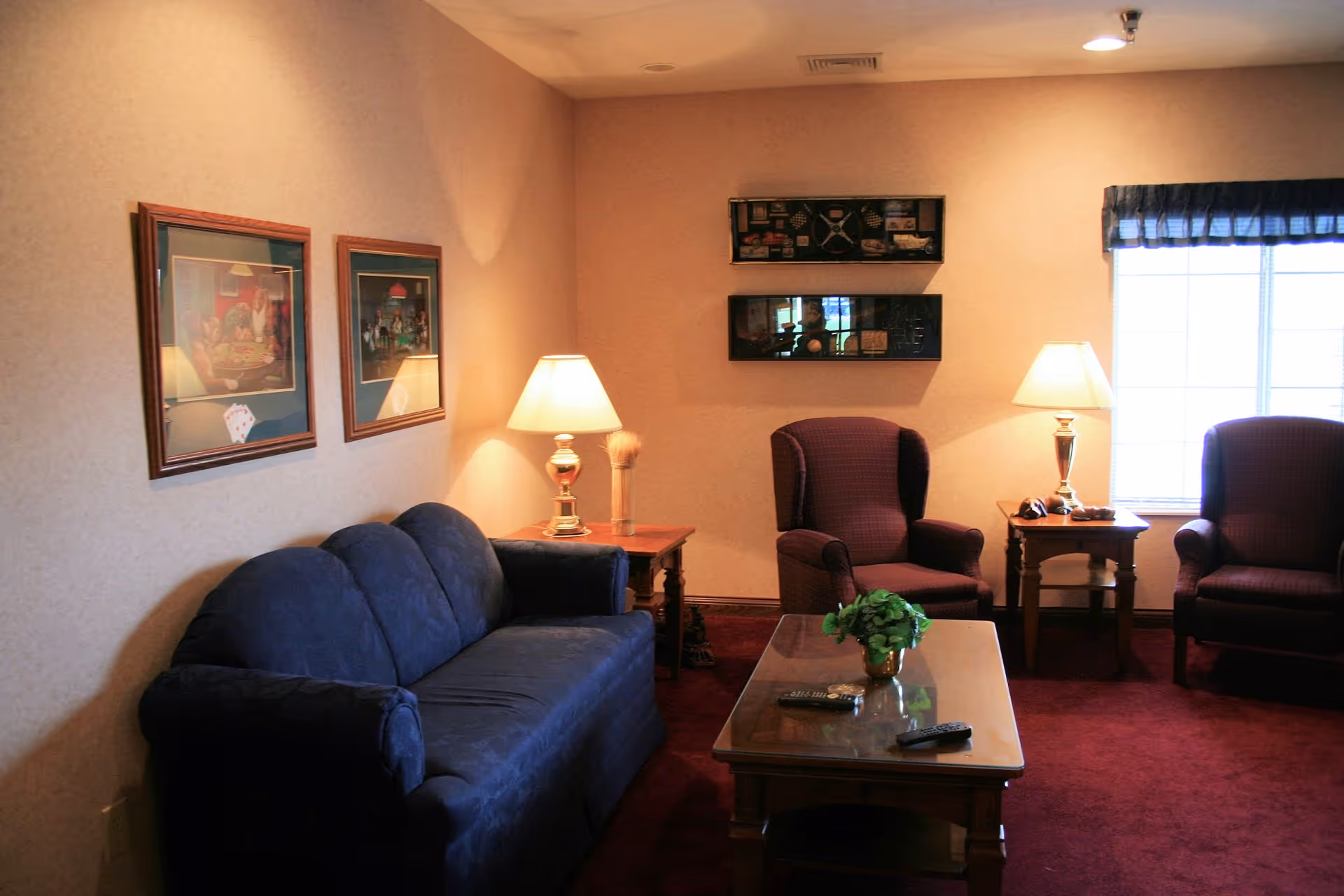 A cozy living room with a dark blue couch on the left, two burgundy armchairs on the right, a wooden coffee table with a small green plant and two remote controls, two side tables each with a lamp, framed pictures on the walls, and a window with a dark valance letting in natural light.