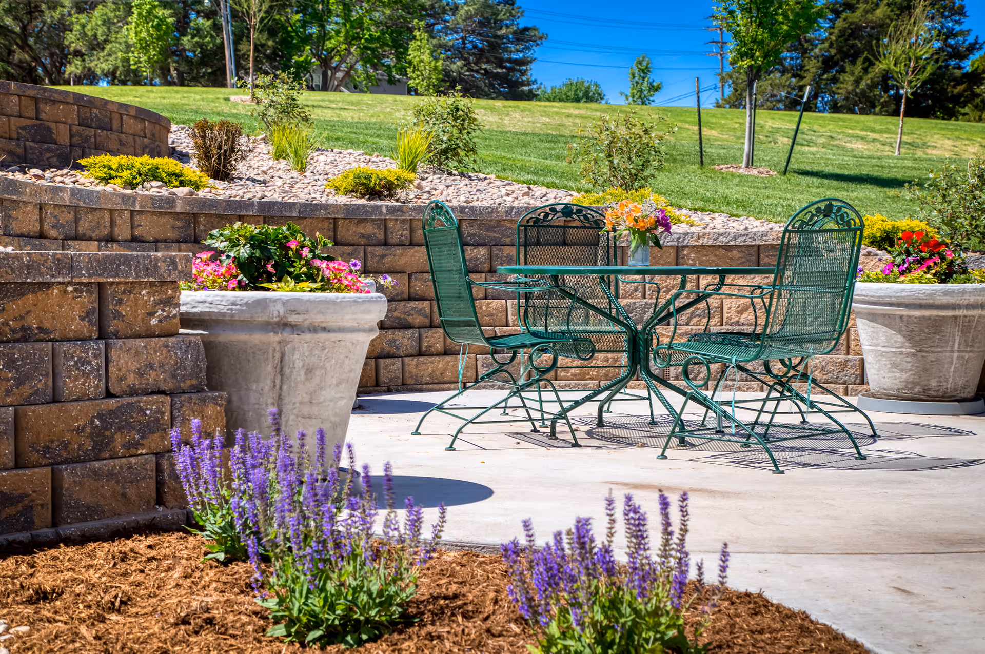 Outdoor patio area with a green metal table and four matching chairs on a concrete surface. The patio is surrounded by large stone retaining walls and large planters filled with colorful flowers. In the foreground, there are purple flowering plants and mulch. The background shows a grassy hill with small trees and a clear blue sky.