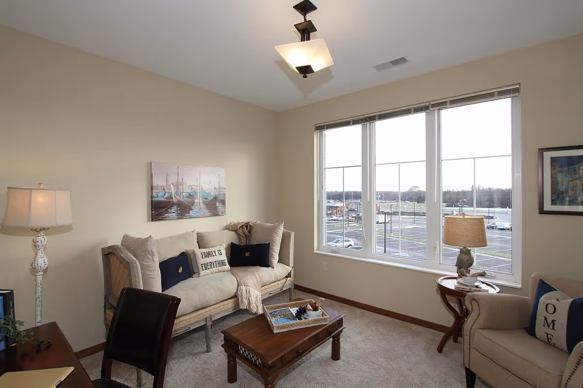 A cozy living room with a beige sofa adorned with cushions, including one that says 'FAMILY IS EVERYTHING.' There is a wooden coffee table with a decorative tray, a beige armchair with a cushion, two table lamps, a floor lamp, and large windows showing a parking lot outside. The walls are light-colored with framed artwork.