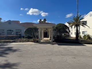 Exterior view of a senior living facility named Ansley Cove showing the entrance with a covered driveway, palm trees, and a clear blue sky with a few clouds.