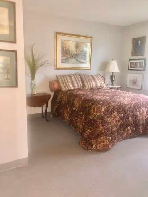 A bedroom with a bed covered in a floral patterned bedspread and several pillows. There is a small wooden side table with a vase containing tall green plants, and a lamp on another table next to the bed. The walls are decorated with framed paintings and photographs.