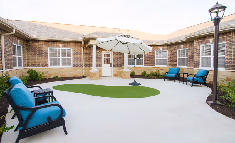 Outdoor courtyard area of a senior living facility with brick walls and multiple windows. The space features a small artificial putting green in the center, surrounded by a concrete patio. There are four black metal chairs with blue cushions arranged around the patio, and a large white umbrella providing shade. A tall lamp post is visible on the right side, and some small plants and shrubs line the edges of the courtyard.