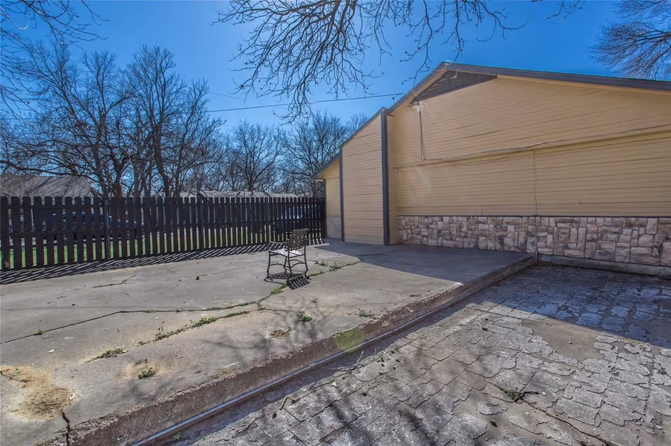 Concrete patio area with a single metal chair beside a yellow building and wooden fence under a clear blue sky.