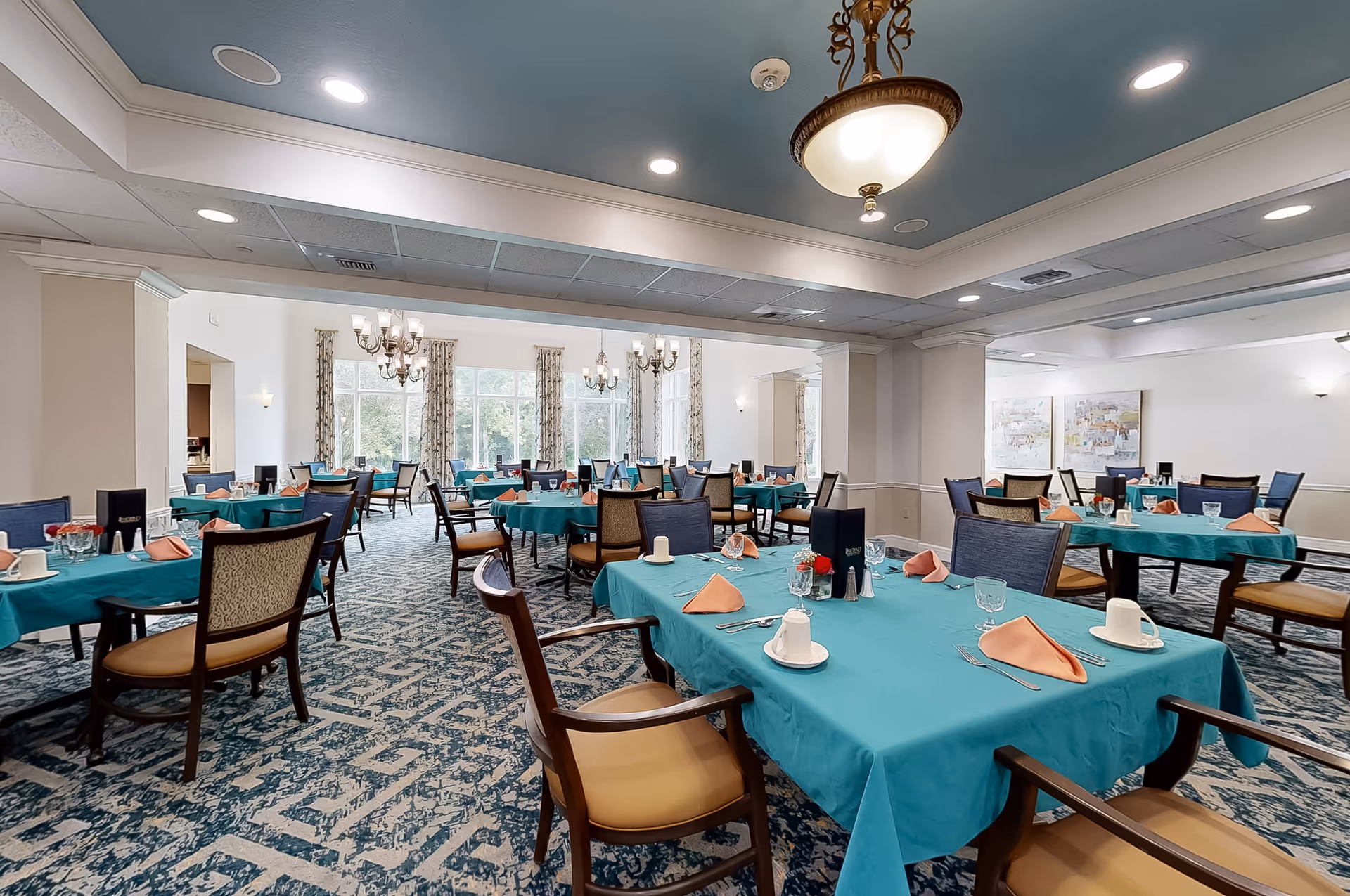 A spacious dining room with multiple tables covered in teal tablecloths, each set with peach-colored napkins, white cups, glasses, and silverware. The room features patterned carpet, large windows with curtains letting in natural light, chandeliers, and comfortable wooden chairs with cushioned seats.