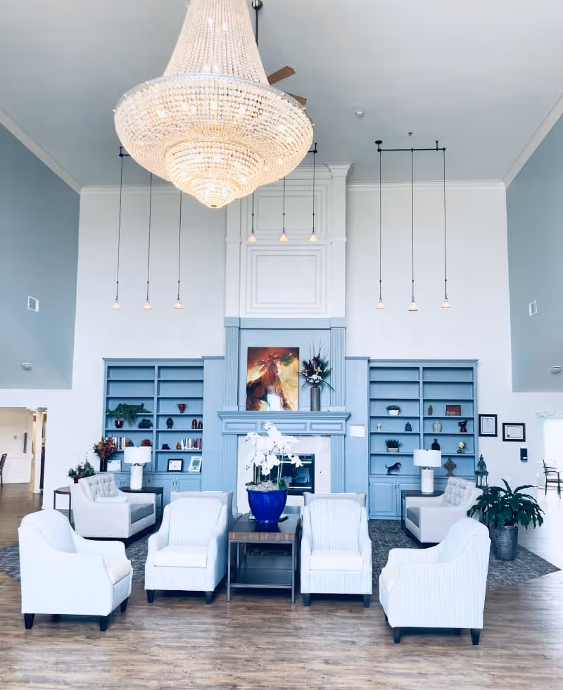 A bright and spacious living room area with a large chandelier hanging from the ceiling. The room features a fireplace with blue built-in shelves on either side, decorated with plants, books, and ornaments. There are four white armchairs arranged around a small wooden table with a blue pot containing white flowers. Additional seating and plants are visible in the background.