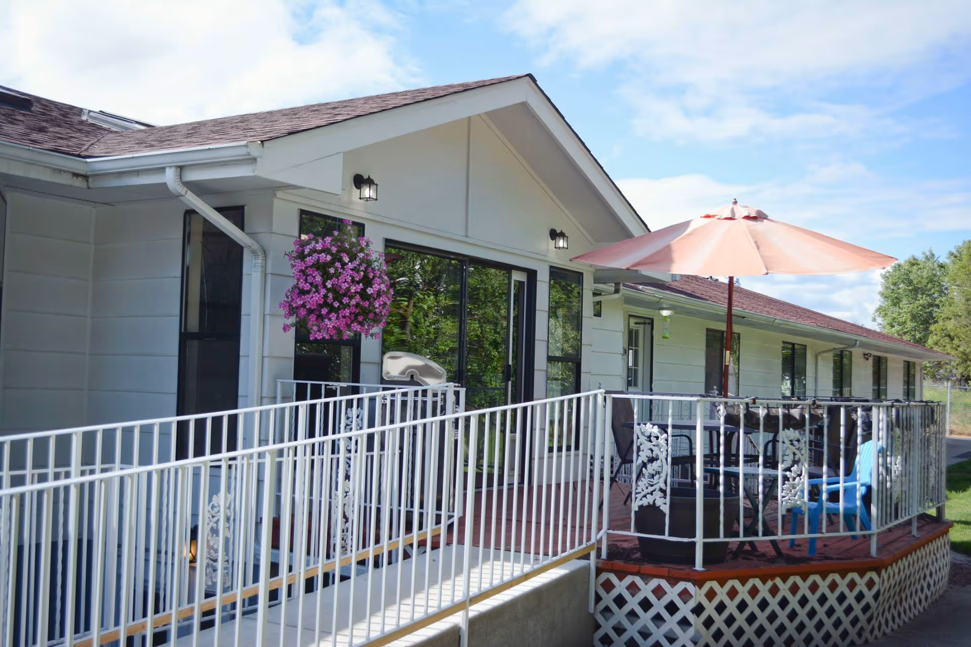 Exterior view of a single-story senior living facility showing a ramped entrance, white railing, a patio with table and umbrella, and hanging flowers.
