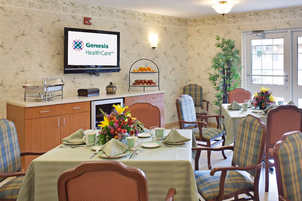 A dining room with tables covered in light green tablecloths, set with plates, cups, utensils, and folded napkins. Each table has a colorful floral centerpiece. The room has wooden chairs with plaid and solid upholstery. A sideboard with a chafing dish, fruit stand, and decorative items is against the wall. A flat-screen TV mounted on the wall displays the Genesis HealthCare logo. There is a large plant near glass doors letting in natural light.