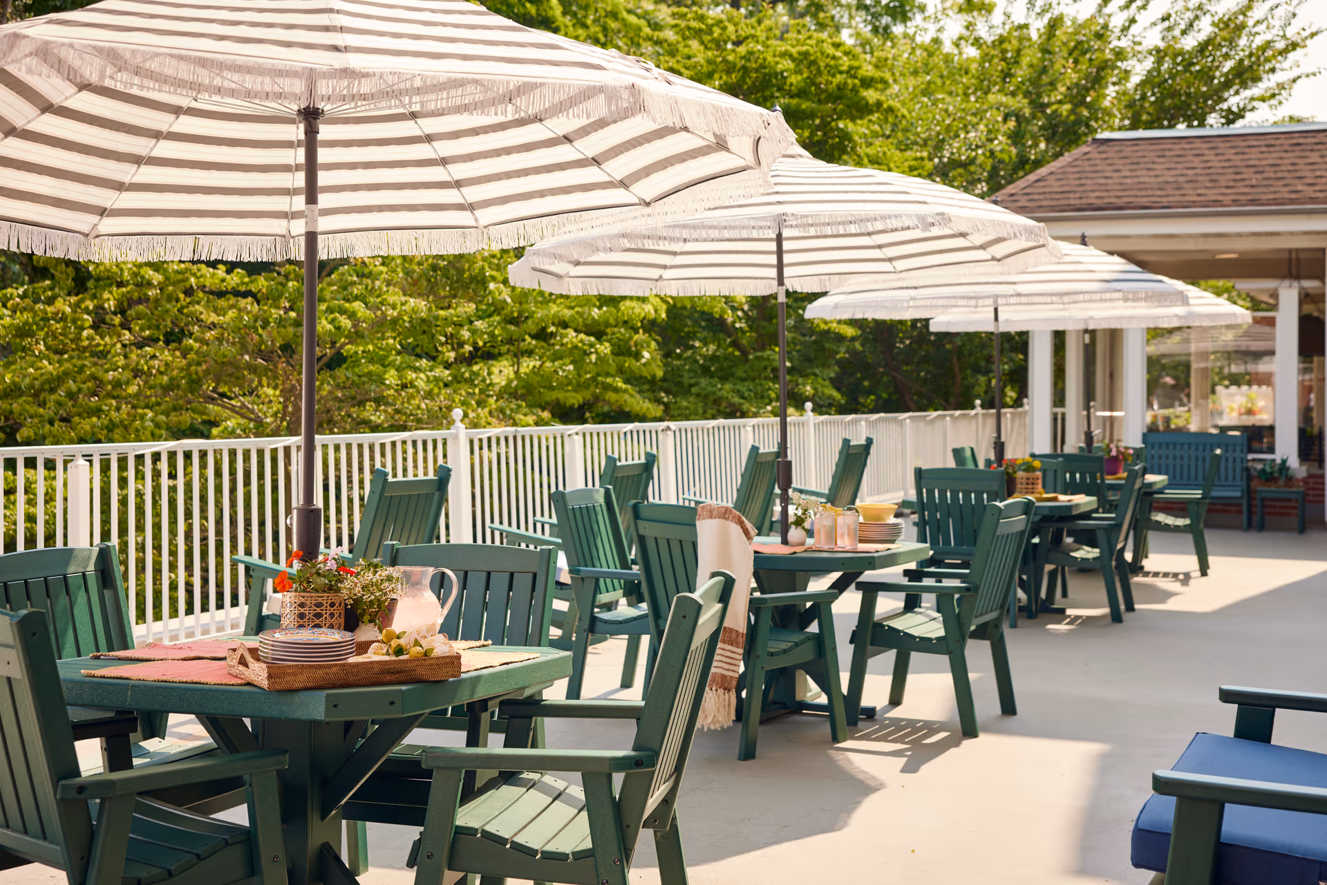 Outdoor patio area with multiple green wooden tables and chairs arranged under large white and beige striped umbrellas. The tables are set with plates, pitchers, and decorative plants. The patio is surrounded by a white railing and lush green trees in the background.