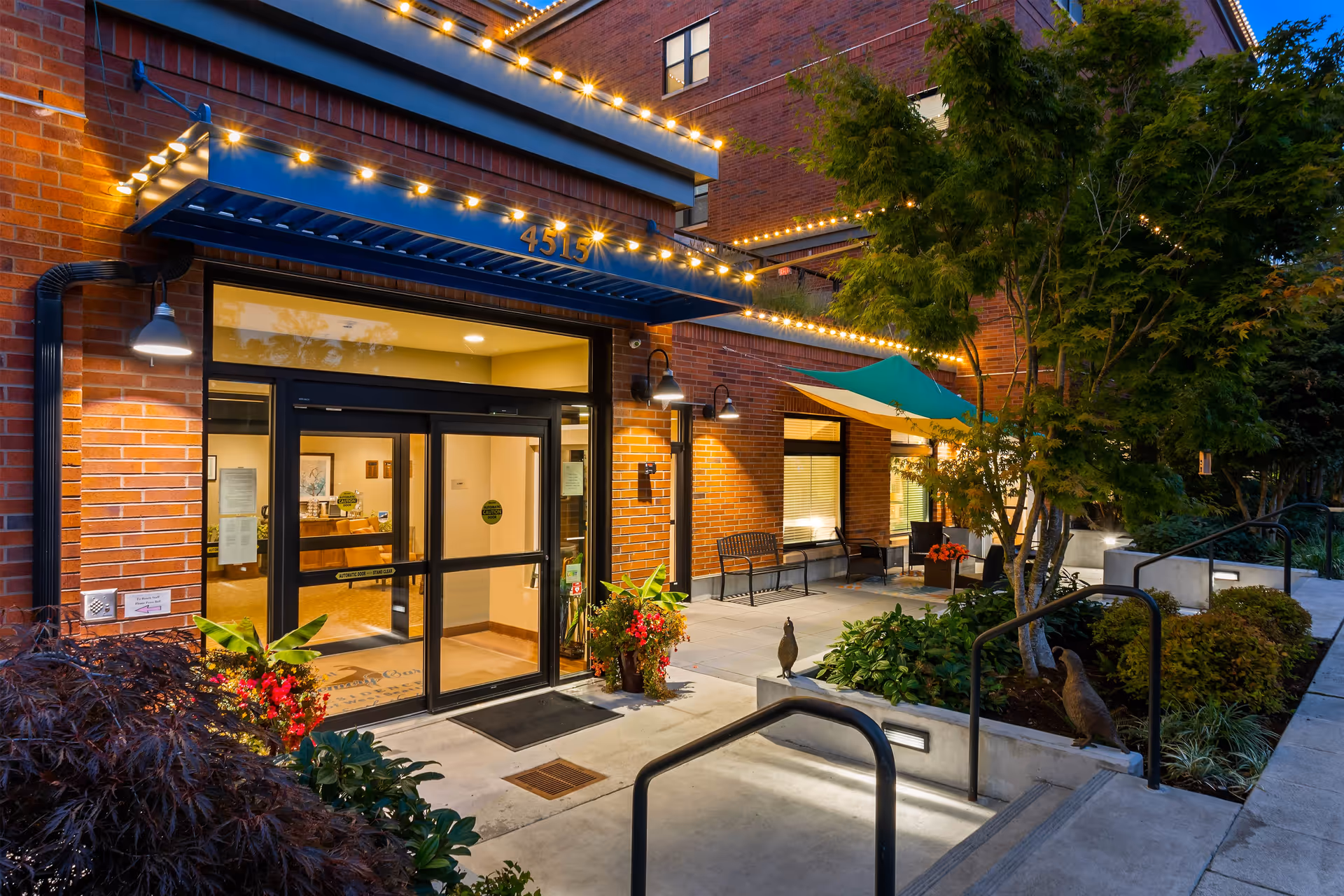Entrance of Quail Park of West Seattle at dusk, featuring a well-lit doorway with glass double doors, brick exterior walls, decorative string lights, potted plants, benches, a green and yellow canopy, and landscaped greenery including trees and shrubs.