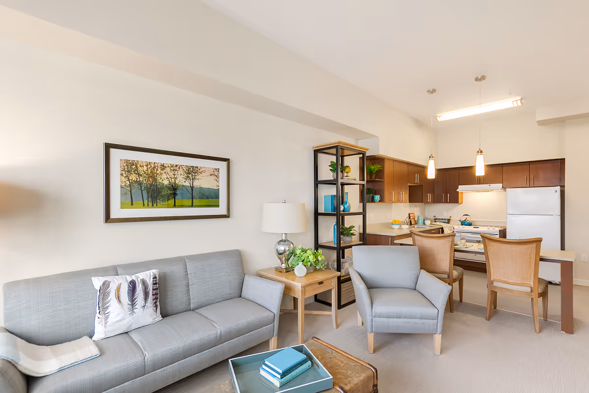 A bright and modern living room and kitchen area in a senior living facility. The living room features a light gray sofa with a decorative pillow, a matching armchair, a wooden side table with a lamp and plant, and a framed landscape painting on the wall. The kitchen has wooden cabinets, a white refrigerator, stove, and two pendant lights hanging over a breakfast bar with two wicker chairs. A black metal and wood shelving unit separates the living room and kitchen spaces.