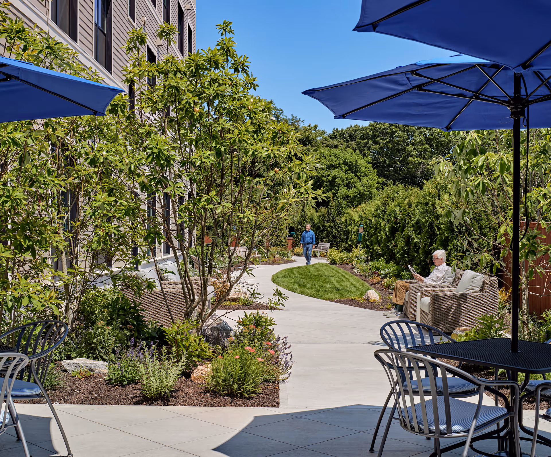 Outdoor patio area at Waterstone on High Ridge with metal tables and chairs under blue umbrellas, a paved walkway surrounded by green shrubs and plants, a man walking in the background, and an elderly person sitting on a wicker sofa reading.